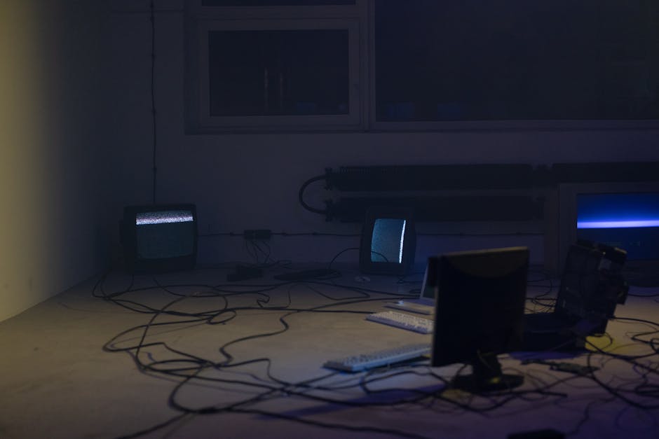 Vintage computer setup with CRT monitors, keyboards, and tangled wires in a dimly lit room.